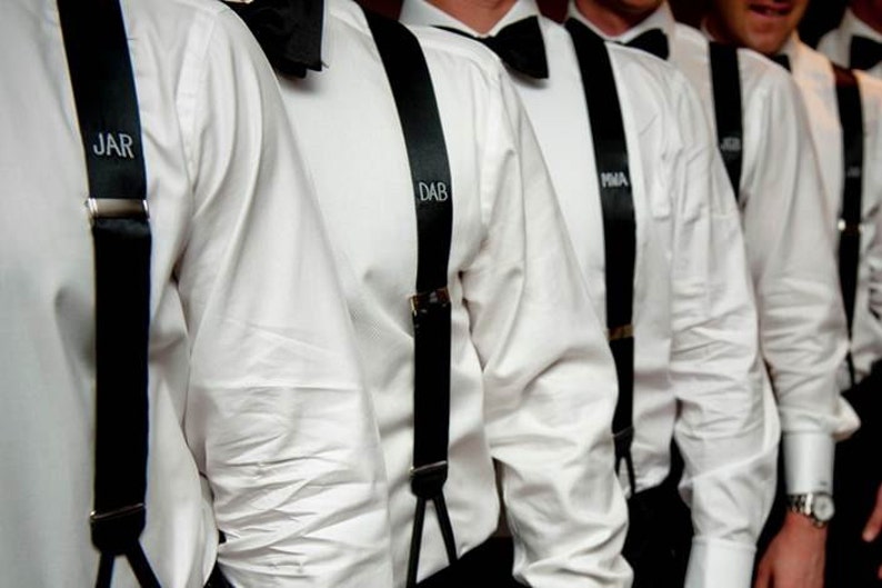 May include: A group of men wearing white shirts with black bow ties and black suspenders. The suspenders have personalized initials on them, including "JAR", "DAB", and "MIA".