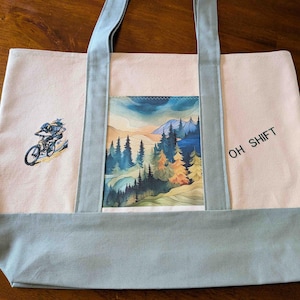 May include: Cream-colored tote bag with light blue accents and handles. Features a mountain biking graphic and a landscape scene with trees and mountains. The text "OH SHIFT" is printed on the side.