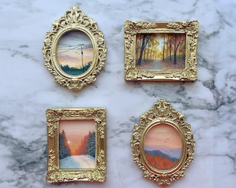 May include: Four miniature paintings in ornate gold-colored frames. Two are oval-shaped and two are rectangular. The paintings depict various landscapes including a sunset with power lines, a forest path, a snowy scene, and a mountain view.
