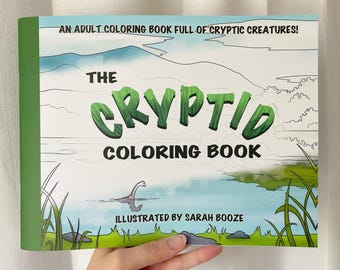 May include: A landscape-oriented coloring book titled The Cryptid Coloring Book with the subtitle An Adult Coloring Book Full of Cryptic Creatures! The cover features a green, textured title, a lake scene with a long-necked creature, and text stating Illustrated by Sarah Booze.