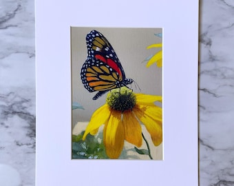 May include: A framed print featuring a monarch butterfly perched on a bright yellow flower. The butterfly has orange, black, and white markings. The flower has yellow petals and a green center. The print is set in a white mat.