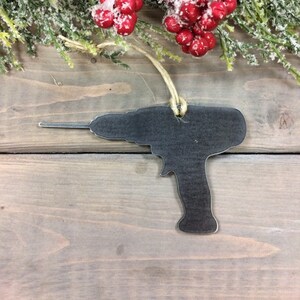 Könnte beinhalten: Ein schwarzer Weihnachtsbaumschmuck aus Metall in Form einer Bohrmaschine. Der Schmuck hängt an einer Schnur aus Bindfaden und befindet sich vor einem Holzgrund.
