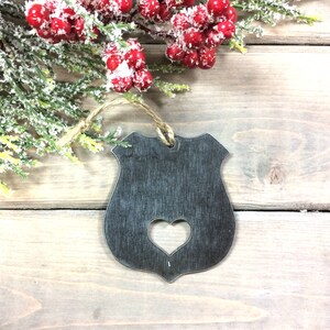 K&ouml;nnte beinhalten: Ein Weihnachtsbaumschmuck aus Metall in Form eines Polizeibadgens mit einem ausgeschnittenen Herzen in der Mitte. Der Anh&auml;nger h&auml;ngt an einer Bindfadenkordel und befindet sich vor einem Holzgrund.