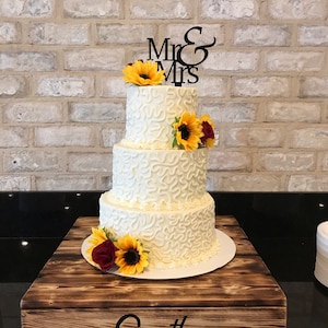 Könnte beinhalten: Eine dreistöckige weiße Hochzeitstorte, verziert mit Buttercreme-Frosting und Sonnenblumen. Die Torte steht auf einem rustikalen Holzkuchenständer mit dem eingravierten Namen "Smith". Der Tortendekor lautet "Mr. & Mrs."