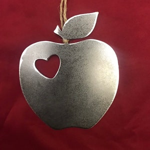 Könnte beinhalten: Ein silberner Apfel-Anhänger aus Metall mit einem ausgeschnittenen Herzen in der Mitte. Der Anhänger hängt an einer Schnur aus Jute.
