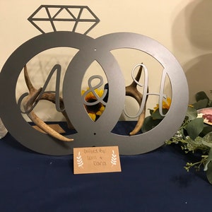 Könnte beinhalten: Ein metallenes Hochzeitsring-Schild mit den Buchstaben A und H in der Mitte, verziert mit Geweih und einem Sonnenblumen-Design. Das Schild steht auf einem blauen Tisch mit einem Schild, auf dem "Geschenk von: Will & Daria" steht.
