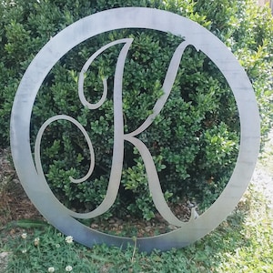 Puede incluir: Un círculo de metal con una letra K decorativa y cursiva recortada en el centro. El círculo es plateado y la letra está sobre un fondo de follaje verde.