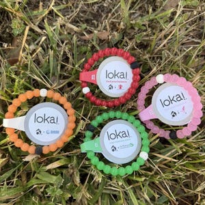May include: Four Lokai bracelets in different colors: red, orange, green, and pink. Each bracelet has a black bead and a white bead. The bracelets have the word "loka!" and the phrase "find your balance" printed on them.