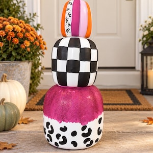 May include: A stacked pumpkin decoration consisting of three tiers. The top pumpkin features vertical stripes in orange, pink, and white with black squiggles. The middle pumpkin has a black and white checkerboard pattern. The bottom pumpkin is white with a pink glitter top and black leopard print spots at the base.