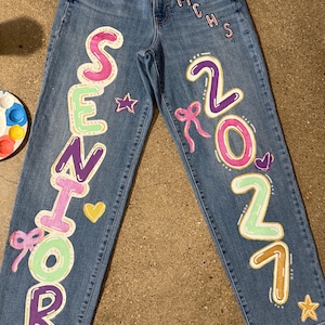 May include: Blue denim jeans featuring hand-painted designs in pink, purple, yellow, and light green. The left leg displays the word SENIOR vertically, while the right leg shows 2027 and MCHS. Decorative elements include stars, hearts, and bows.
