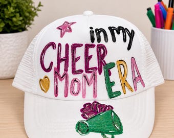 Könnte beinhalten: Eine weiße Trucker-Cap mit dem Schriftzug "in my CHEER MOM ERA" in verschiedenen Farben wie Schwarz, Lila, Grün und Gelb. Das Design umfasst einen rosa Stern, ein gelbes Herz und eine grüne Megafon-Grafik auf der Vorderseite.