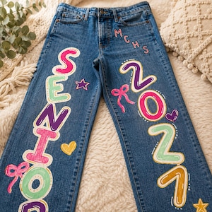 May include: Blue denim jeans featuring hand-painted designs. The left leg displays the word SENIOR in colorful bubble letters with pink bows and a yellow heart. The right leg shows 2027, MCHS, a pink bow, a purple heart, and yellow stars.