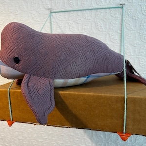 May include: Plush purple whale toy with a geometric pattern, resting on a brown cardboard box. The whale has small black eyes and fins. The box is suspended by green string.