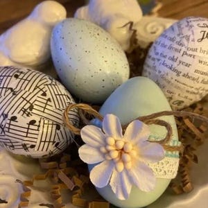 Op de afbeelding: Een verzameling decoratieve paaseieren in een wit keramisch schaaltje. De eieren zijn beschilderd in verschillende kleuren, waaronder lichtblauw en wit, en versierd met muzieknoten, tekst en bloemmotieven. Decoratieve vogels zijn op de achtergrond.