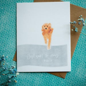 May include: Greeting card with a watercolor illustration of a golden retriever puppy. The card has the text "Just want to say hello" in a handwritten font. The card is on a brown envelope, with a teal patterned background.