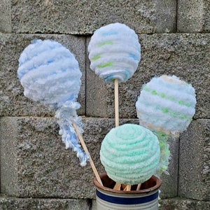 May include: Four yarn balls on sticks in a white ceramic vase with a blue stripe. The yarn balls are blue, green, and white.