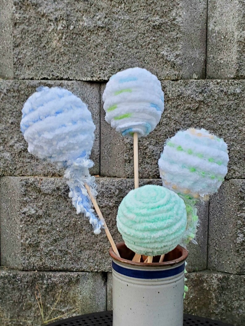 May include: Four yarn balls on sticks in a white ceramic vase with a blue stripe. The yarn balls are blue, green, and white.