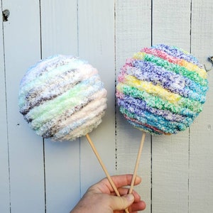 May include: Two yarn balls on sticks, one is rainbow colored and the other is a mix of white, brown, and green.