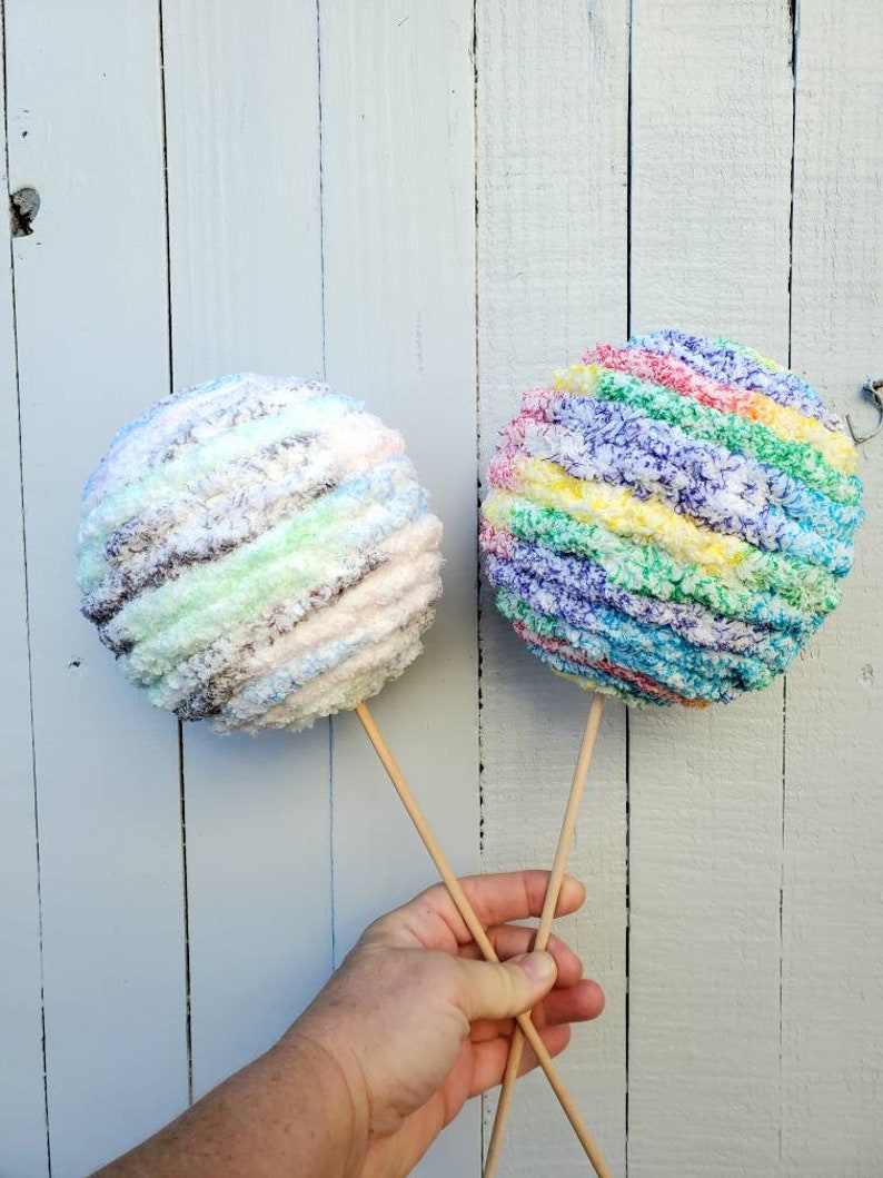 May include: Two yarn balls on sticks, one is rainbow colored and the other is a mix of white, brown, and green.