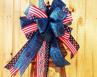 May include: A decorative bow featuring American flag patterns with red, white, and blue stripes and stars. The blue ribbon sections include text such as HERO, PEACE MAKERS, BRAVE, and PROTECT & SERVE, along with police badge and shield graphics.