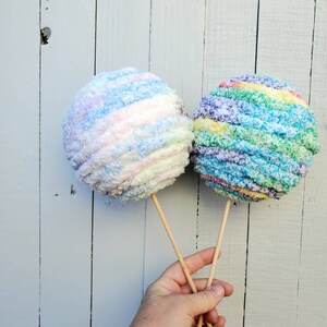 May include: Two colorful yarn balls on sticks, one is blue, white, and pink, the other is green, blue, and purple.