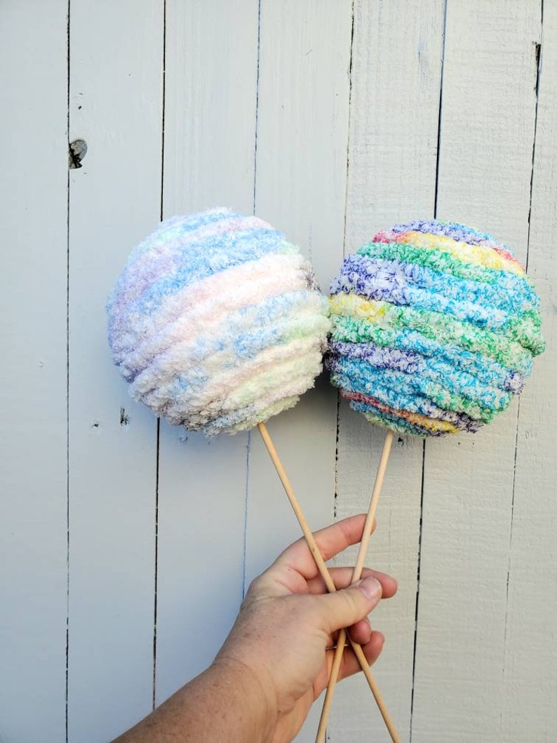 May include: Two colorful yarn balls on sticks, one is blue, white, and pink, the other is green, blue, and purple.