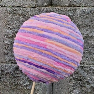 May include: A large, round, pink, purple, and orange yarn ball on a wooden stick. The yarn ball is striped and fluffy.