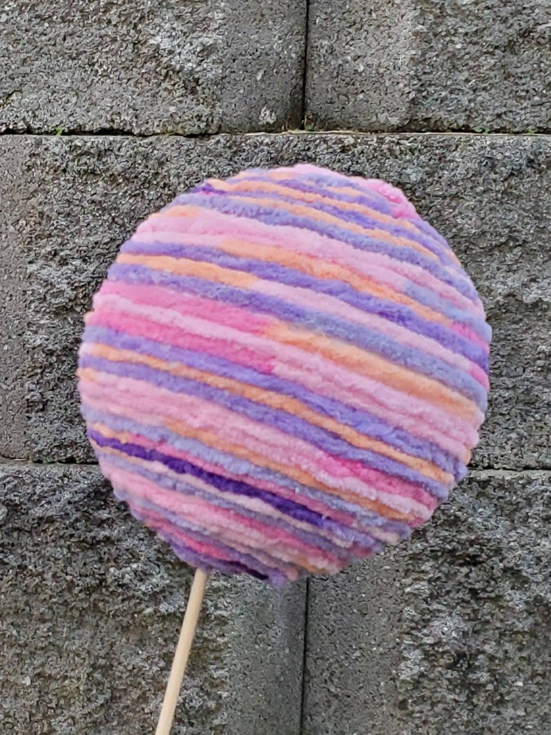 May include: A large, round, pink, purple, and orange yarn ball on a wooden stick. The yarn ball is striped and fluffy.