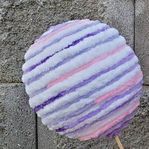 May include: A large, round, striped ball made of yarn in shades of white, pink, and purple. The ball is fluffy and soft, and it looks like it would be fun to touch.
