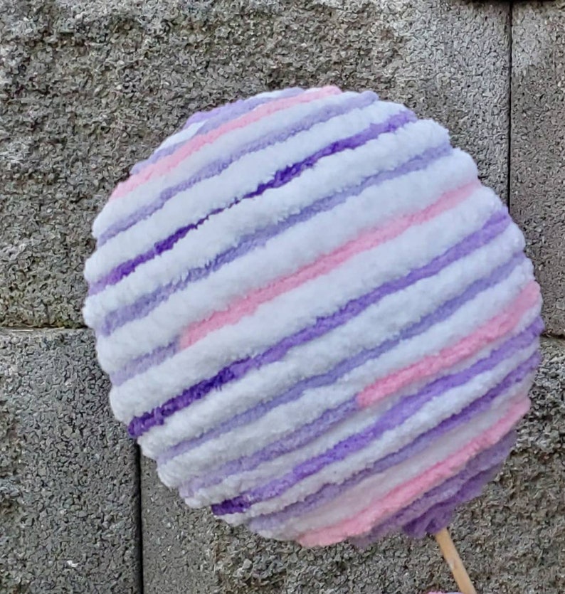 May include: A large, round, striped ball made of yarn in shades of white, pink, and purple. The ball is fluffy and soft, and it looks like it would be fun to touch.