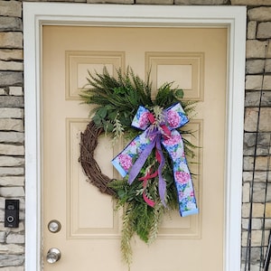 May include: A wreath with a floral print ribbon bow, featuring pink and blue hydrangeas, on a brown door with a white frame.