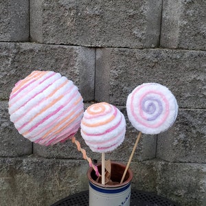 May include: Three large, colorful, striped and swirl-patterned lollipops on sticks in a white ceramic pot. The lollipops are pink, white, and orange.