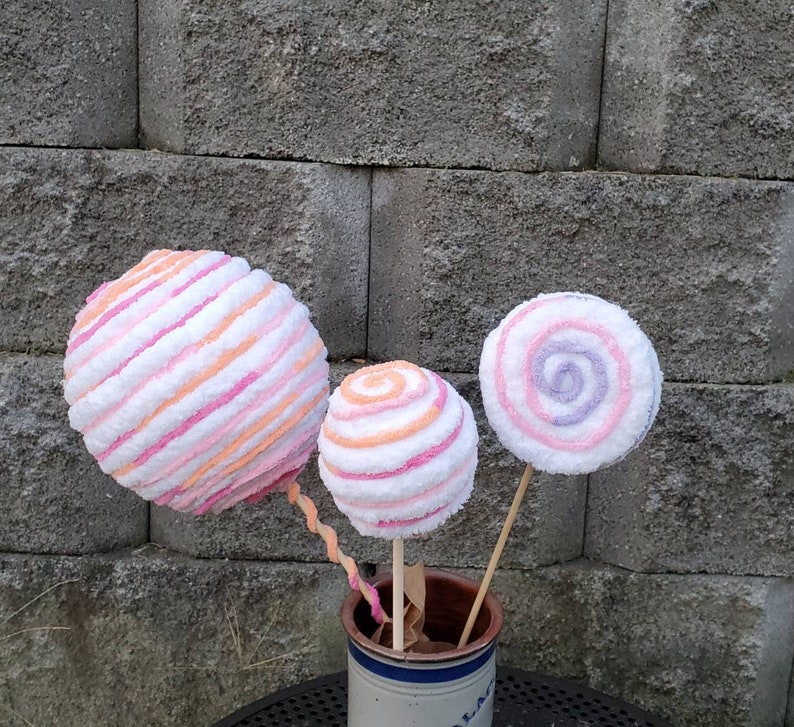May include: Three large, colorful, striped and swirl-patterned lollipops on sticks in a white ceramic pot. The lollipops are pink, white, and orange.