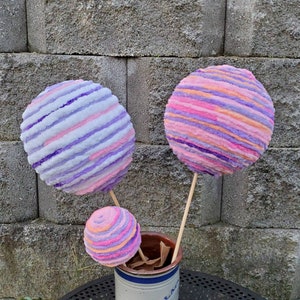 May include: Three yarn balls on sticks, in shades of pink, purple, and white, arranged in a vase. The yarn balls are striped and resemble cotton candy.
