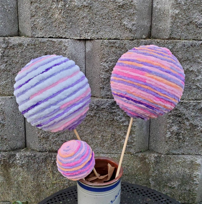 May include: Three yarn balls on sticks, in shades of pink, purple, and white, arranged in a vase. The yarn balls are striped and resemble cotton candy.