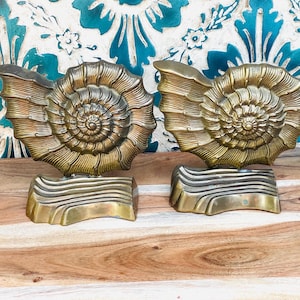 May include: Two antique brass seashell bookends. Each bookend features a detailed spiral design and rests on a rectangular base with horizontal ridges. The bookends are displayed on a wooden surface.