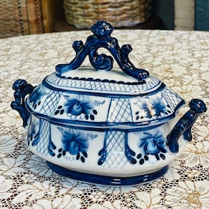May include: A white ceramic sugar bowl with a lid, featuring hand-painted blue floral patterns and a cross-hatched grid design. The bowl has two side handles and a decorative finial on the lid, all accented with deep blue glaze details.