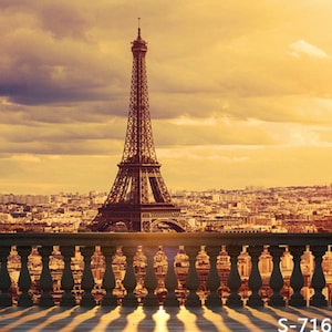 May include: A view of the Eiffel Tower in Paris, France, from a balcony with a stone railing. The sky is a warm orange color, and the sun is setting behind the tower. The city of Paris is visible in the background.