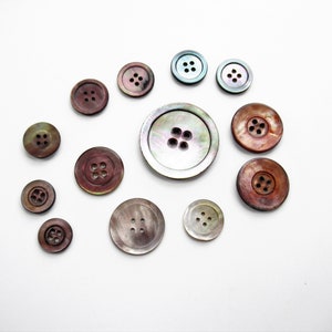 May include: A collection of 12 iridescent mother-of-pearl buttons in various sizes and shades of brown, blue, and green. The buttons have four holes each.