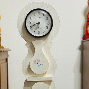 May include: A cream-colored wooden clock with a round black clock face. The clock has a decorative floral design and is standing on a wooden surface. The clock face has black numbers and hands.