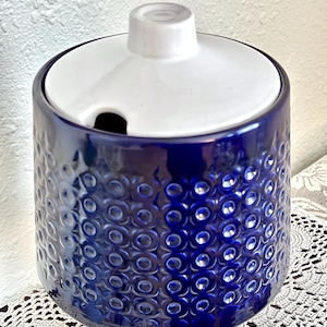 May include: A cobalt blue ceramic jar with a white lid. The jar has a textured pattern of raised circles. The lid has a small knob and a slot for a spoon. The jar is likely for storing sugar or other dry goods.