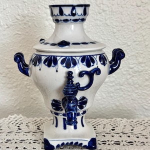 May include: White ceramic samovar with blue floral patterns and accents. The tea kettle features a spigot, handles, and a lid. The design includes a base and a decorative top. A traditional tea brewing vessel.
