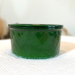 May include: A small, round, emerald green glass bowl. The bowl has a diamond pattern etched into the sides. The rim of the bowl is a darker shade of green. The bowl is sitting on a white, textured surface.