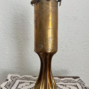 May include: A tall, cylindrical brass vase with a metallic gold finish. The upper section features a textured, stippled surface pattern, while the lower base is smooth and flares outward with vertical, concave fluted indentations.