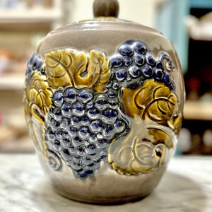 May include: A ceramic jar with a lid, featuring a grape design. The jar is a muted gray color, with a brown lid and a detailed relief of blue grapes and gold leaves. The jar is likely used for storage.