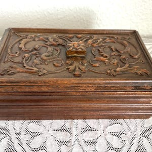 May include: A rectangular wooden box with a dark brown finish. The lid is intricately carved with floral and scroll designs, and a small handle is centered on the top. The box is likely used for storage or display.