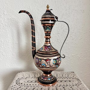 May include: An ornate copper pitcher with a long, curved spout and a handle. The pitcher is decorated with colorful floral patterns and intricate black detailing. The lid is topped with a small gold finial. The pitcher is displayed on a lace doily.