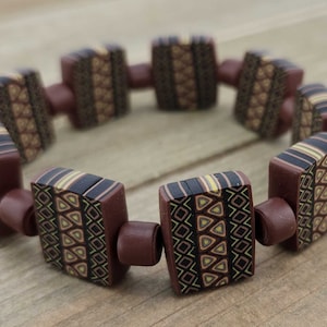 May include: A brown bracelet with square-shaped beads featuring a geometric pattern in black, brown, and gold.
