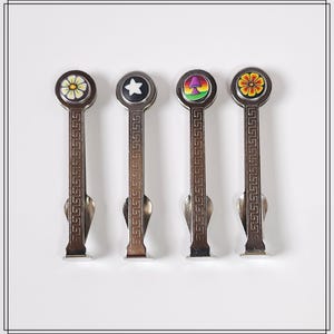 May include: Four metal tea bag holders with decorative tops. Each holder has a different design: a yellow flower, a white star, a mushroom, and a yellow and red flower. The holders have a decorative pattern on the handle.
