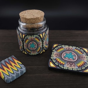 Op de afbeelding: Een kleine glazen pot met een kurken stopper en decoratief polymeerklei-ontwerp. Het ontwerp heeft een centrale turquoise schildpad omgeven door geometrische patronen in geel, zwart en grijs. Twee bijpassende decoratieve tegels zijn ook aanwezig.